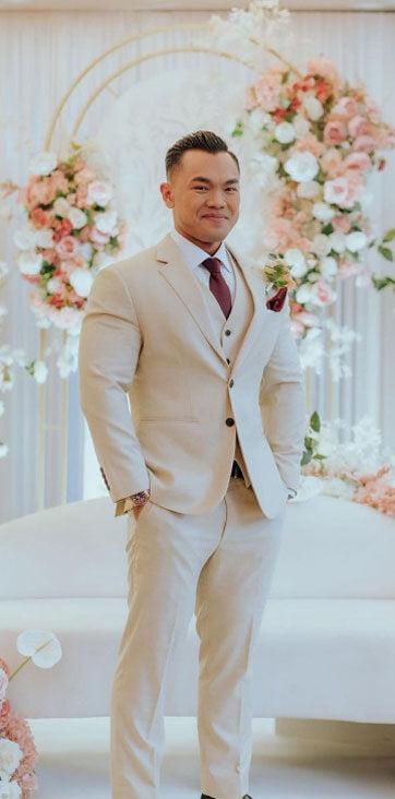 Groom in a custom beige suit with a burgundy tie, standing in front of floral wedding decorations